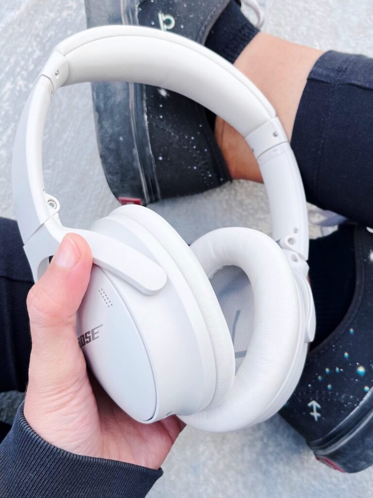 A hand holding white Bose QuietComfort 45 noise-cancelling headphones, recommended for an optimal sound healing and immersive listening experience.