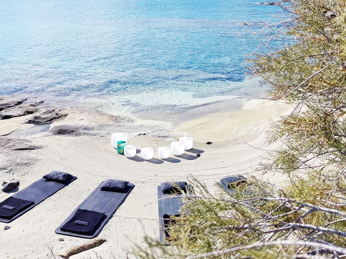 Crystal singing bowls set up by the Aegean Sea on Kimolos, with black meditation mats placed on the sand under a tree.