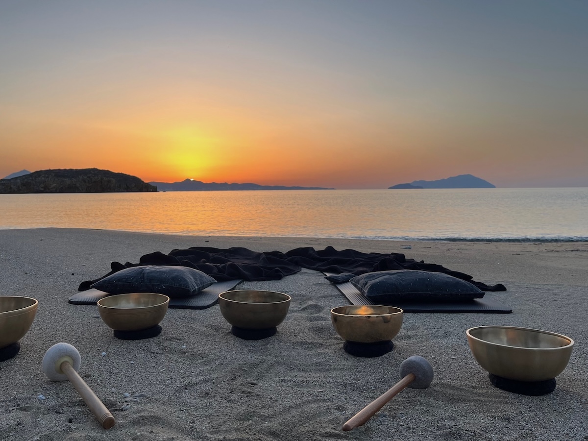 Crystal singing bowls and meditation mats arranged for a sunset Sound Healing on Kimolos beach, with Tibetan bowls in the sand and a golden horizon.