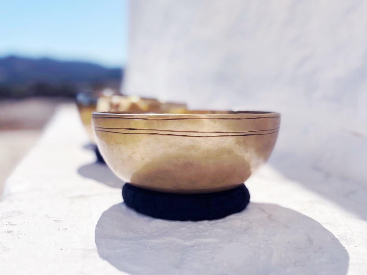 A close-up of three Tibetan singing bowls in the frequencies of Uranus, Jupiter and Mars/Neptune resting on black cushions outdoors against a white wall, often used in a Personalized Sound Bath with those frequencies.
